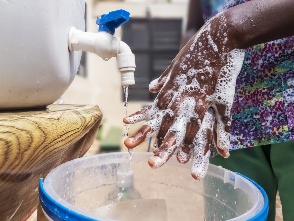 Close-up of hands covered in soap suds being washed under a white plastic tap with a blue handle, water flowing into a clear bucket with a blue rim, against a blurred background of wall and window.