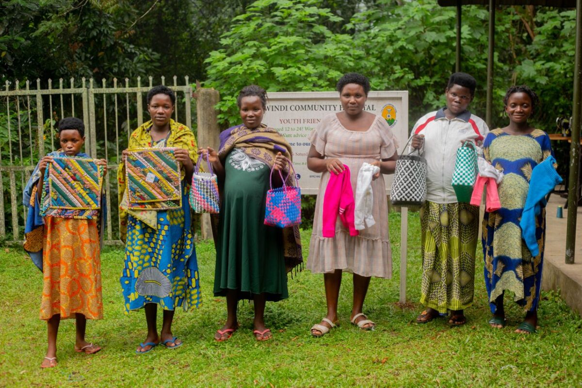 A group of African women showcasing colorful handmade crafts outdoors at Bwindi Community Hospital.