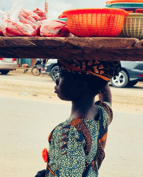 African woman carrying baskets on her head in Nigerian city street market. Traditional attire and street scene.
