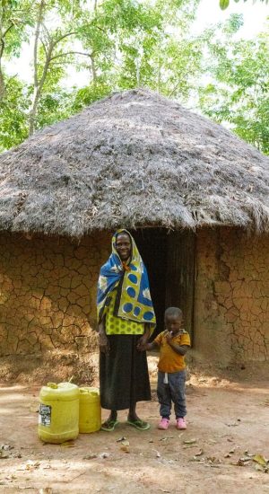 A woman and child stand outside a traditional mud hut with thatched roof in a rural area.