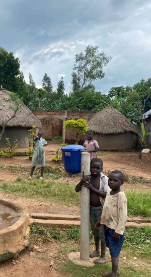 Children standing near a hand‑operated water pump in a rural village with traditional huts, greenery, and cloudy sky in the background.